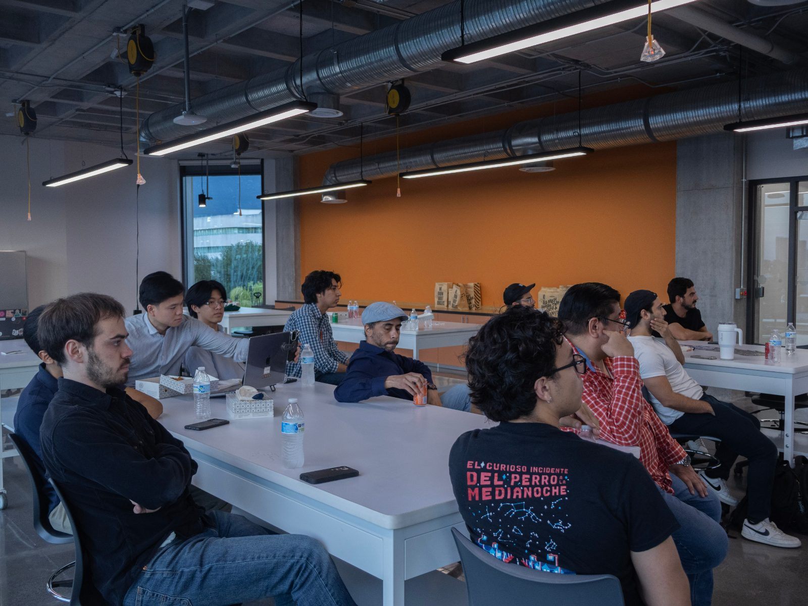 A group seated around a long white table at a meetup, listening to someone speak.